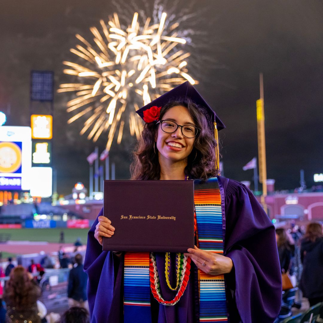 Graduate at Commencement holds up her diploma while fireworks explode at Giants stadium