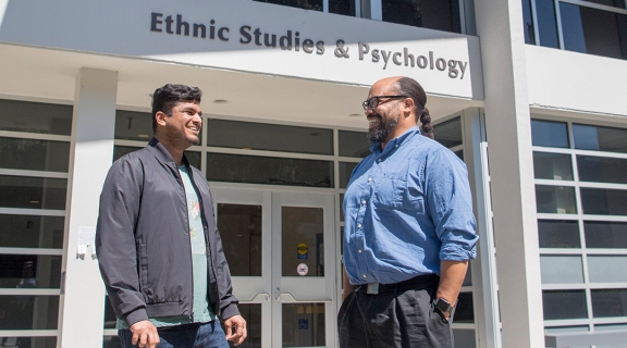 Two men talk in front of the Ethnic Studies and Psychology building on campus